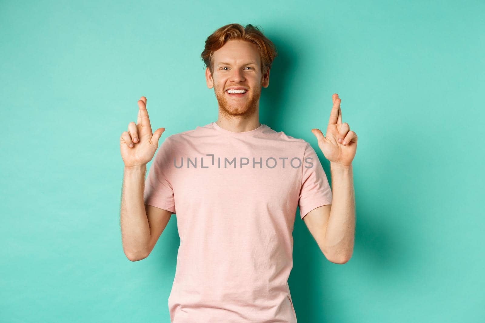Optimistic guy with red hair and beard smiling, cross fingers for good luck and looking hopeful at camera, making a wish, standing over mint background.