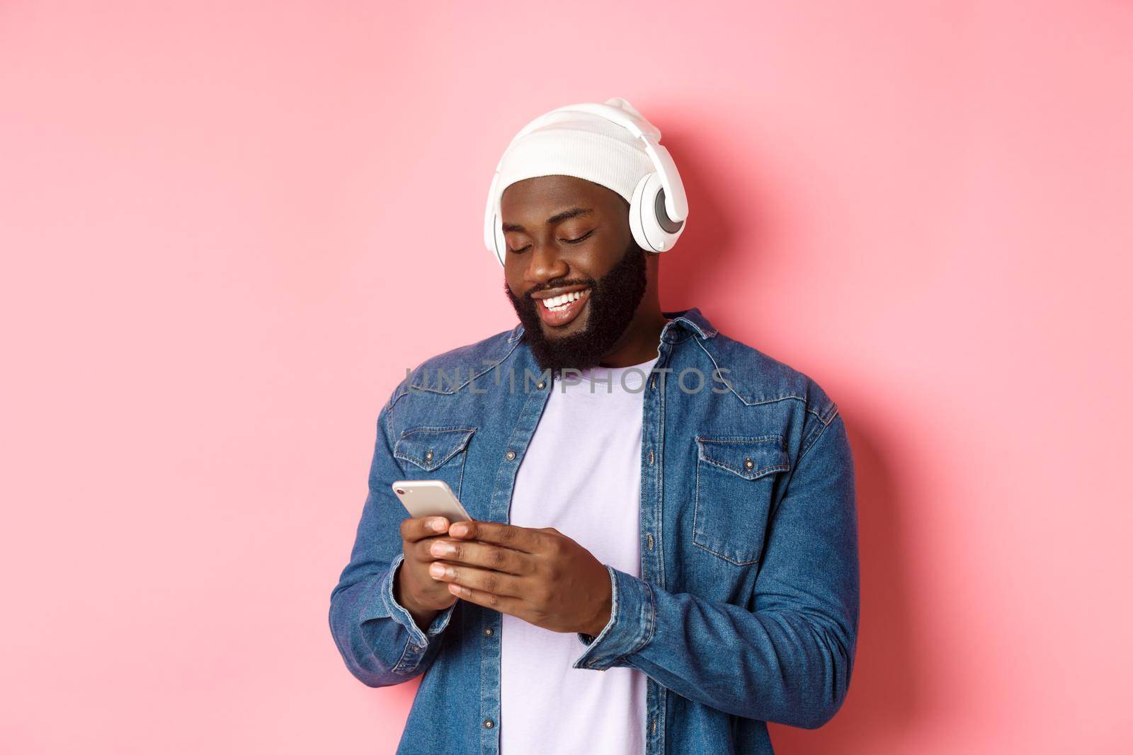 Handsome stylish Black man listening music in headphones and writing message on smartphone, smiling happy, standing over pink background.