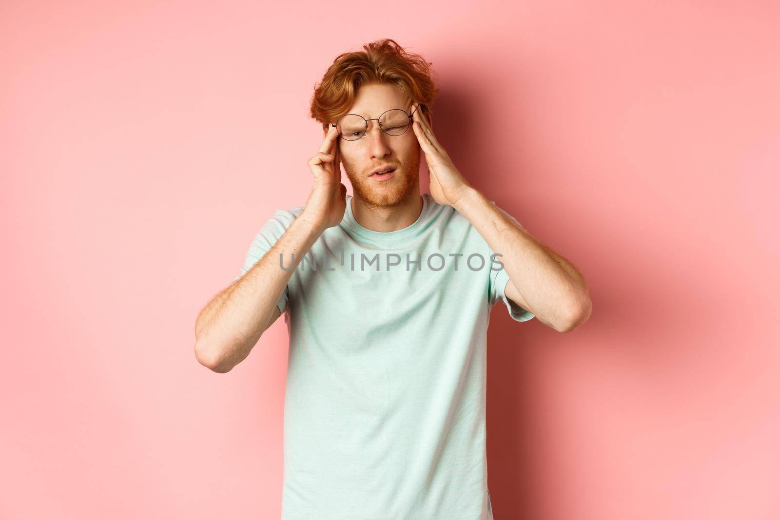 Portrait of redhead man in crooked glasses touching head and feeling dizzy or nauseous, having hangover or headache, standing over pink background.
