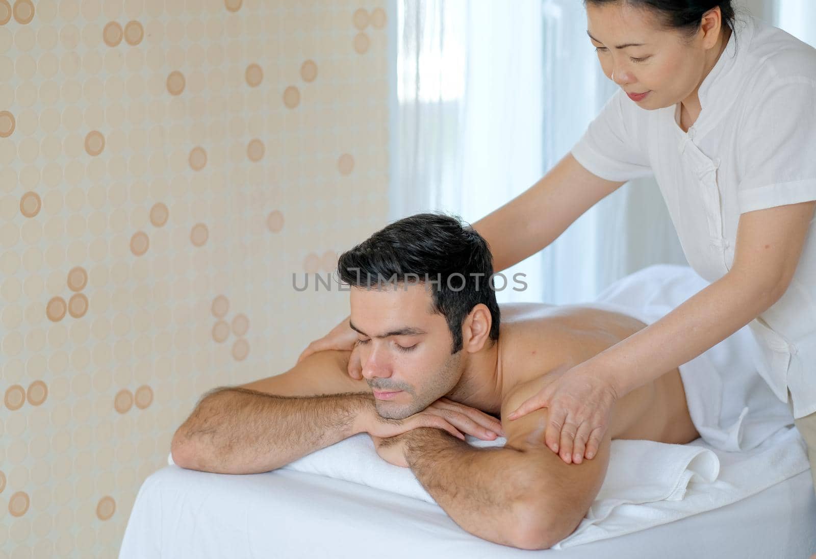 Handsome man enjoy with Thai masseuse massage to him and he lie on bed in spa room with day light.