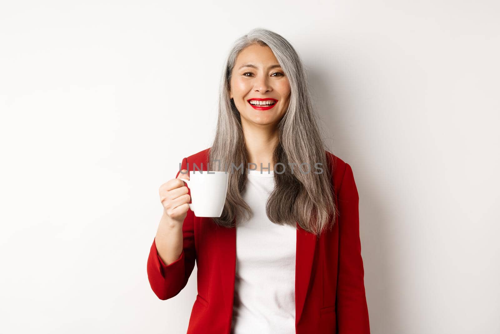 Business people concept. Happy asian businesswoman in red blazer smiling, drinking coffee from mug, standing over white background.