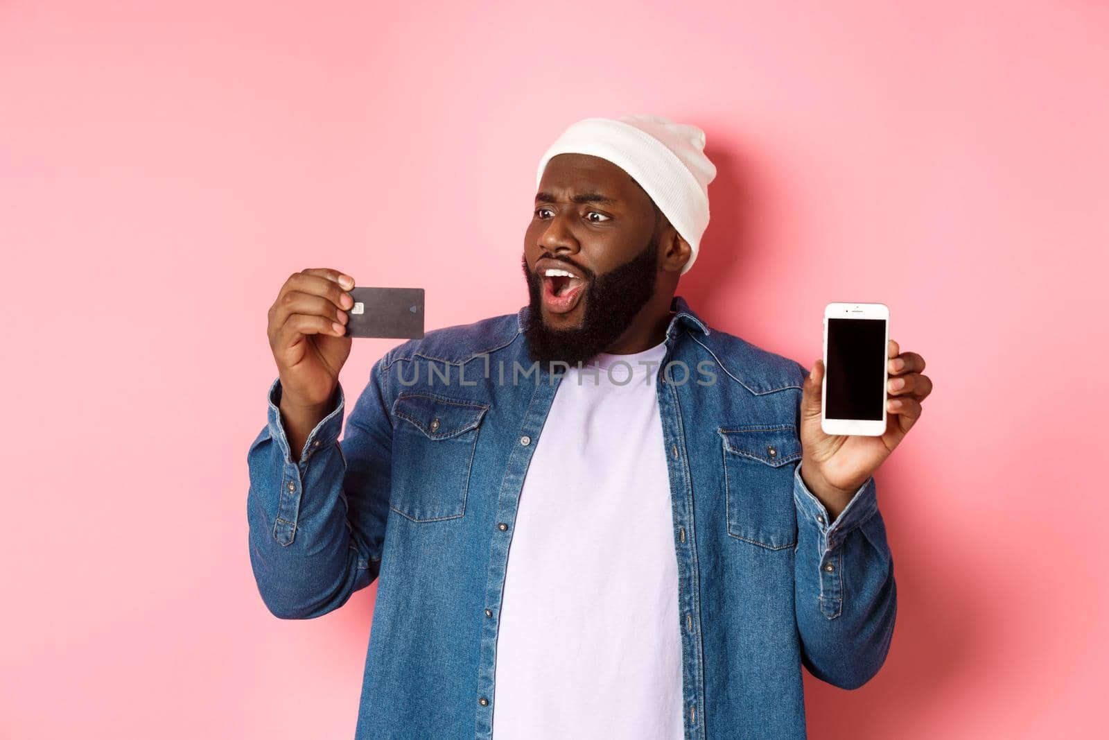 Online shopping. Shocked Black man showing mobile phone screen, looking startled at credit card, standing in hipster clothes against pink background.