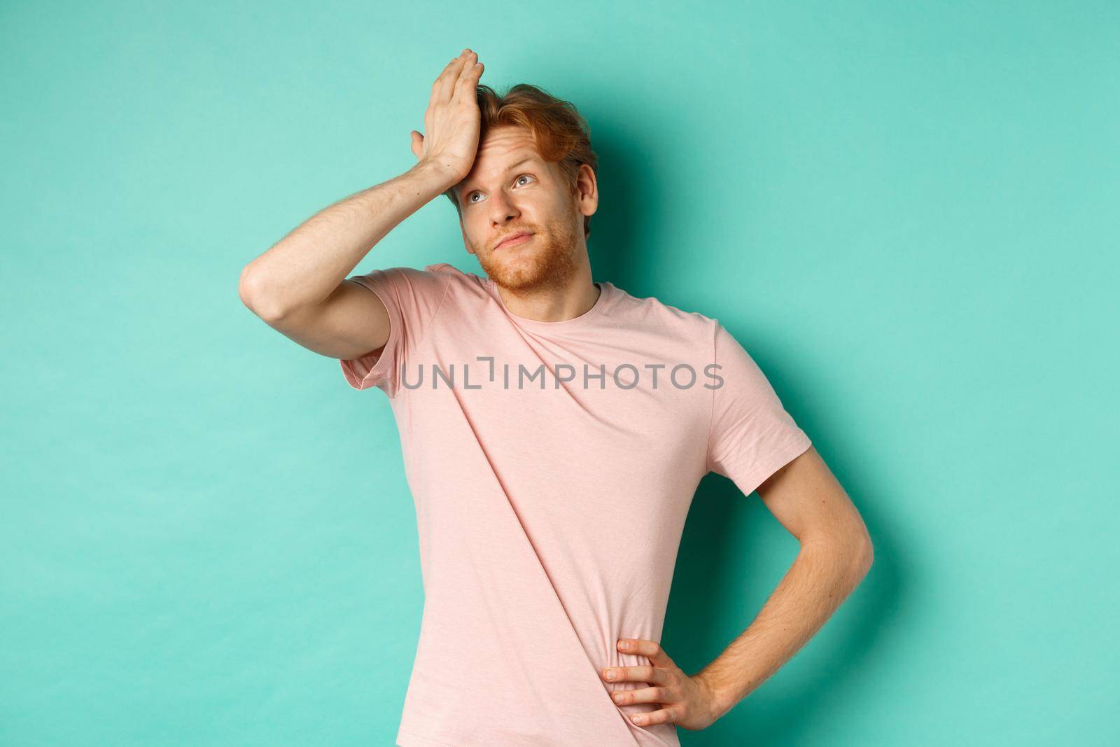 Annoyed and bothered redhead male model showing facepalm and roll eyes, standing over mint background. Copy space