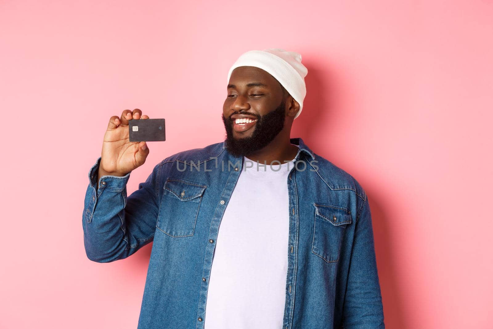 Shopping concept. Image of happy african-american man looking satisfied at credit card, recommending bank, standing over pink background.