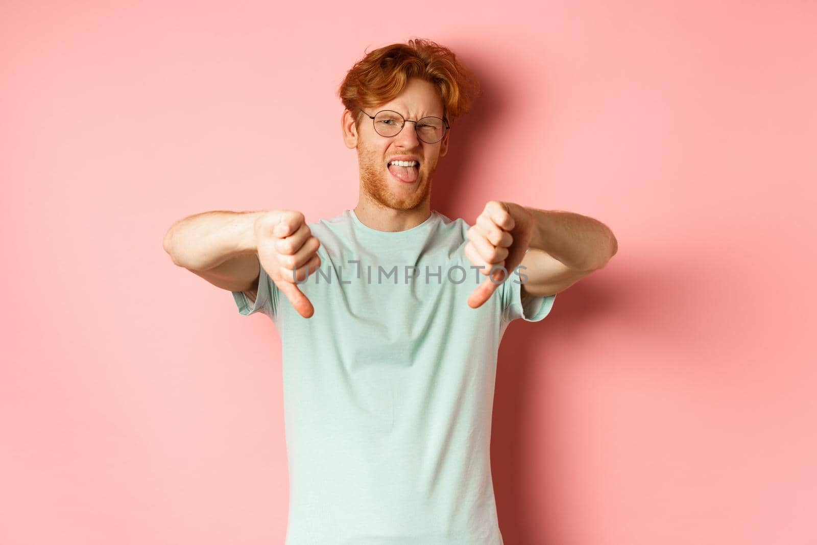 Disgusted redhead man in t-shirt and glasses showing thumbs down and tongue, grimacing from aversion, standing over pink background.