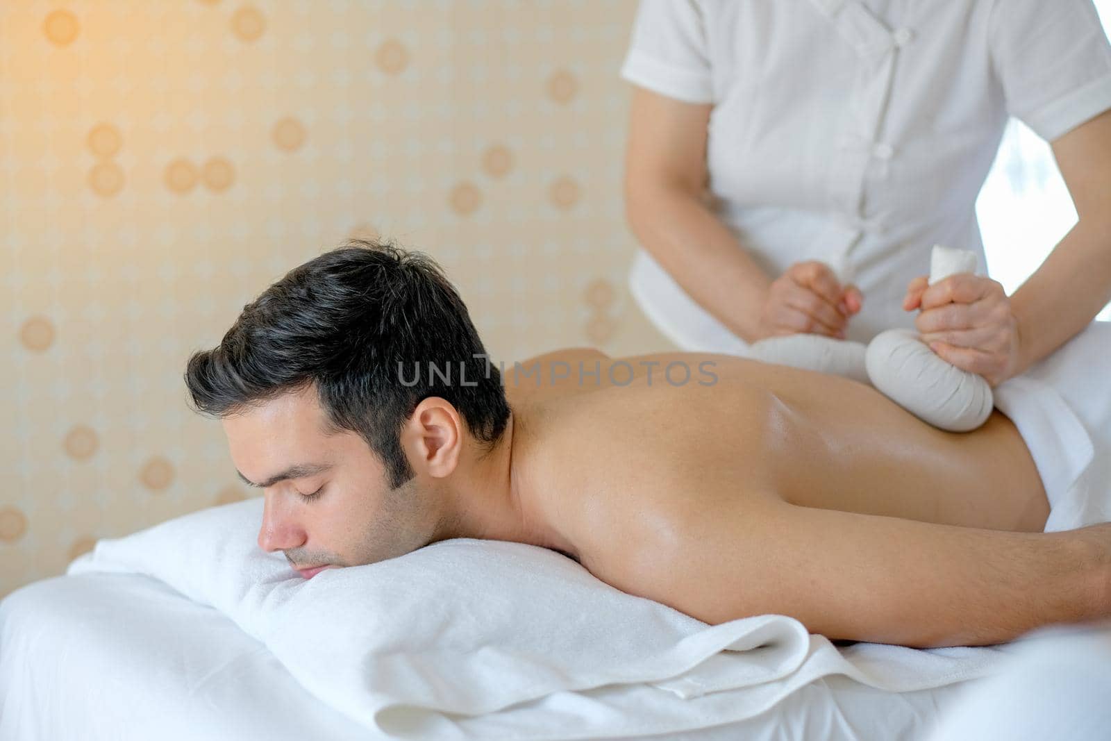 Handsome man enjoying with massage by herbal ball on his back in spa room with day light.