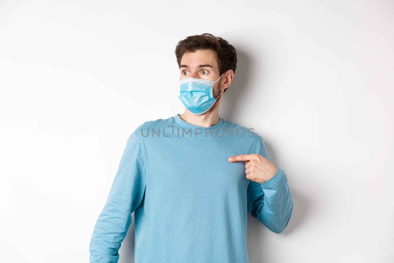 Covid-19, health and quarantine concept. Confused man in medical mask pointing at himself, looking left with ambushed face, standing over white background by Benzoix