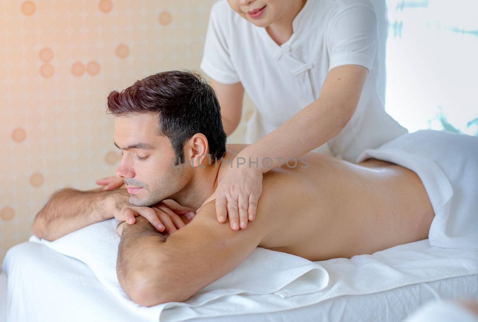 Portrait of handsome man enjoy with Thai masseuse massage to him and he lie on bed in spa room with day light.