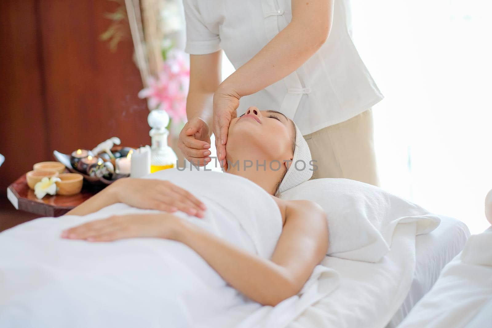 Portrait of Thai masseuse do face massage for  young beautiful girl who lie and look up on bed in spa room with day light.