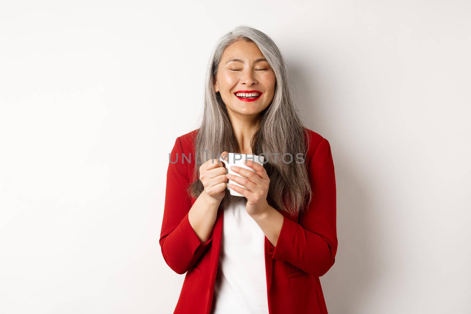 Business people concept. Happy office lady in red blazer enjoying drinking coffee, holding mug and smiling delighted, standing over white background by Benzoix