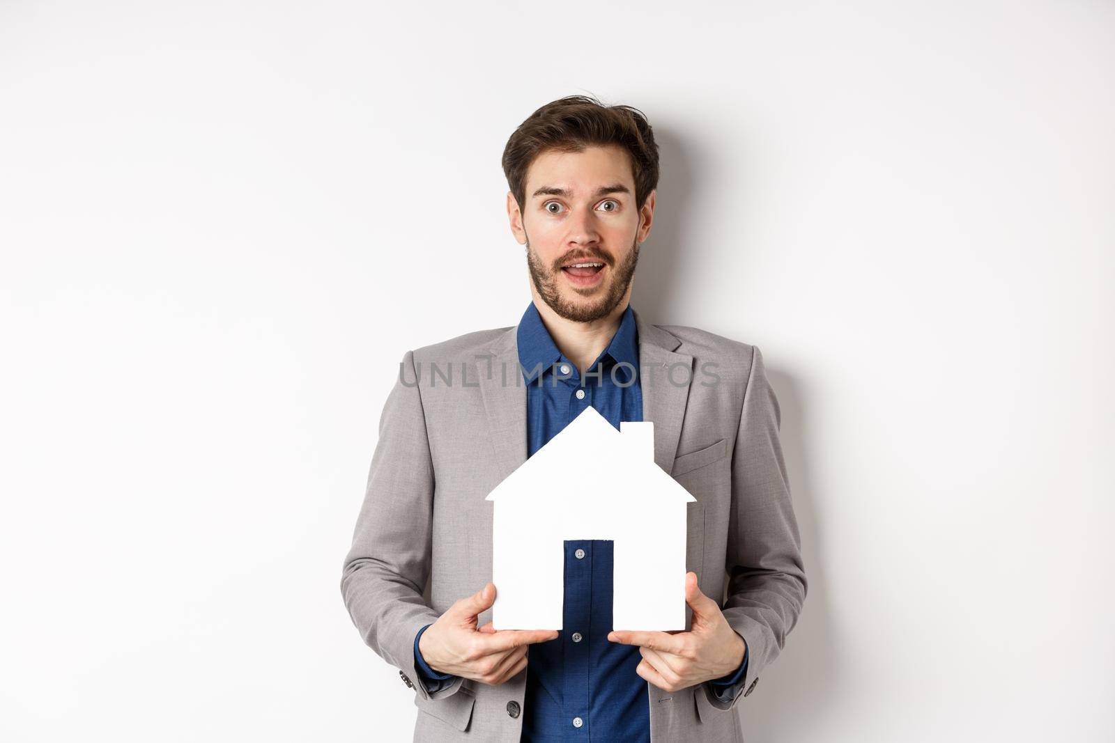 Real estate. Excited business man in suit holding paper house cutout and looking amazed at camera, buying apartment, white background by Benzoix