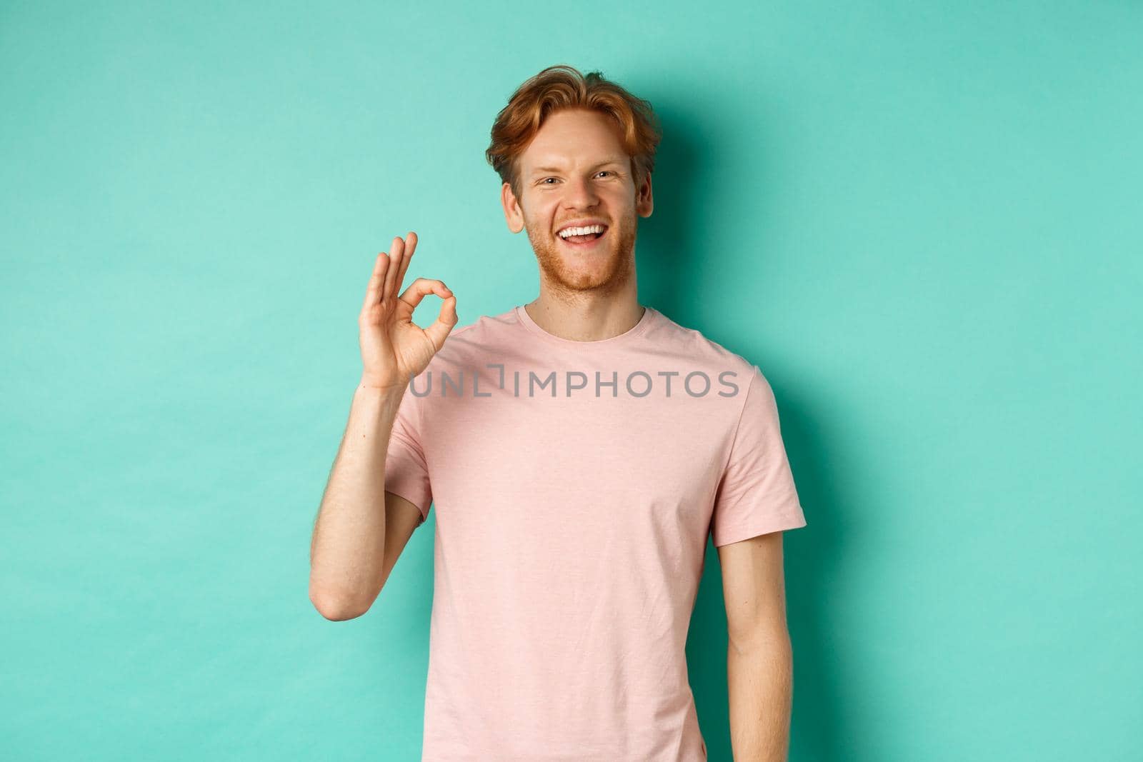 Handsome young bearded man in t-shirt showing Ok sign, smiling with white teeth and saying yes, agree with you, standing over turquoise background.