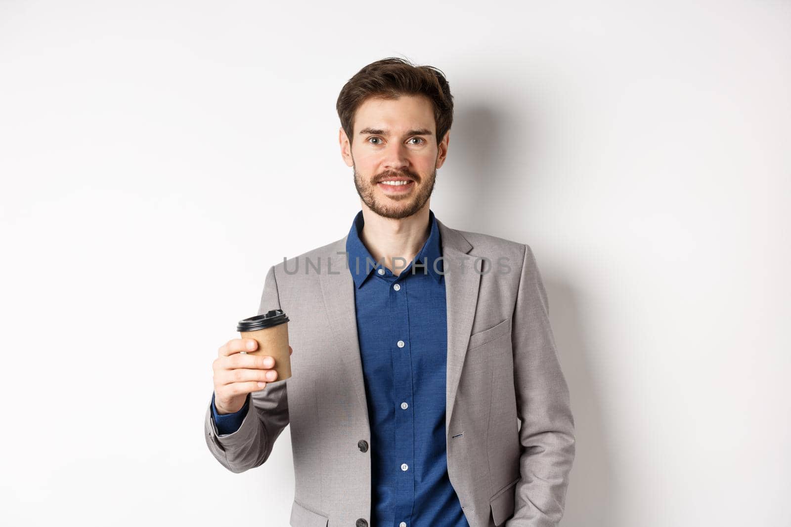 Smiling office worker in suit drinking coffee from paper cup, having break after work at cafe, standing against white background.