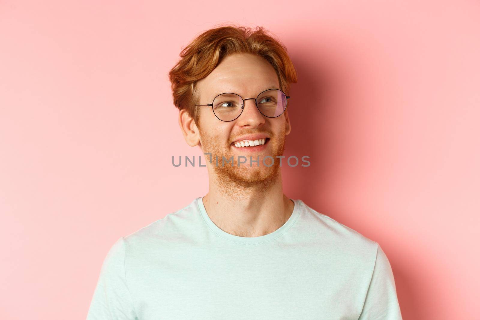 Face of handsome redhead man in glasses smiling pleased, looking at upper right corner, standing over pink background.