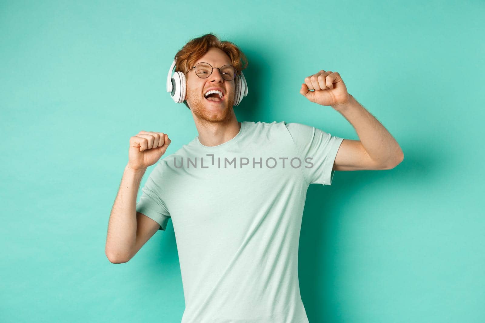 Lifestyle concept. Carefree guy with red hair, dancing and having fun, listening music in headphones, standing over turquoise background.