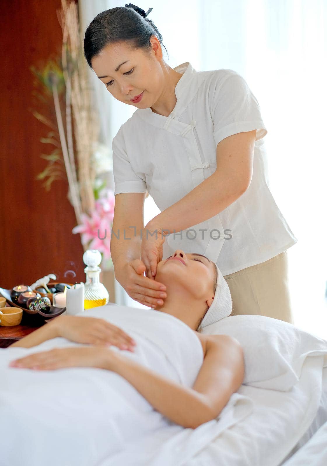 Thai masseuse do face massage for  young beautiful girl who lie and look up on bed in spa room with day light.