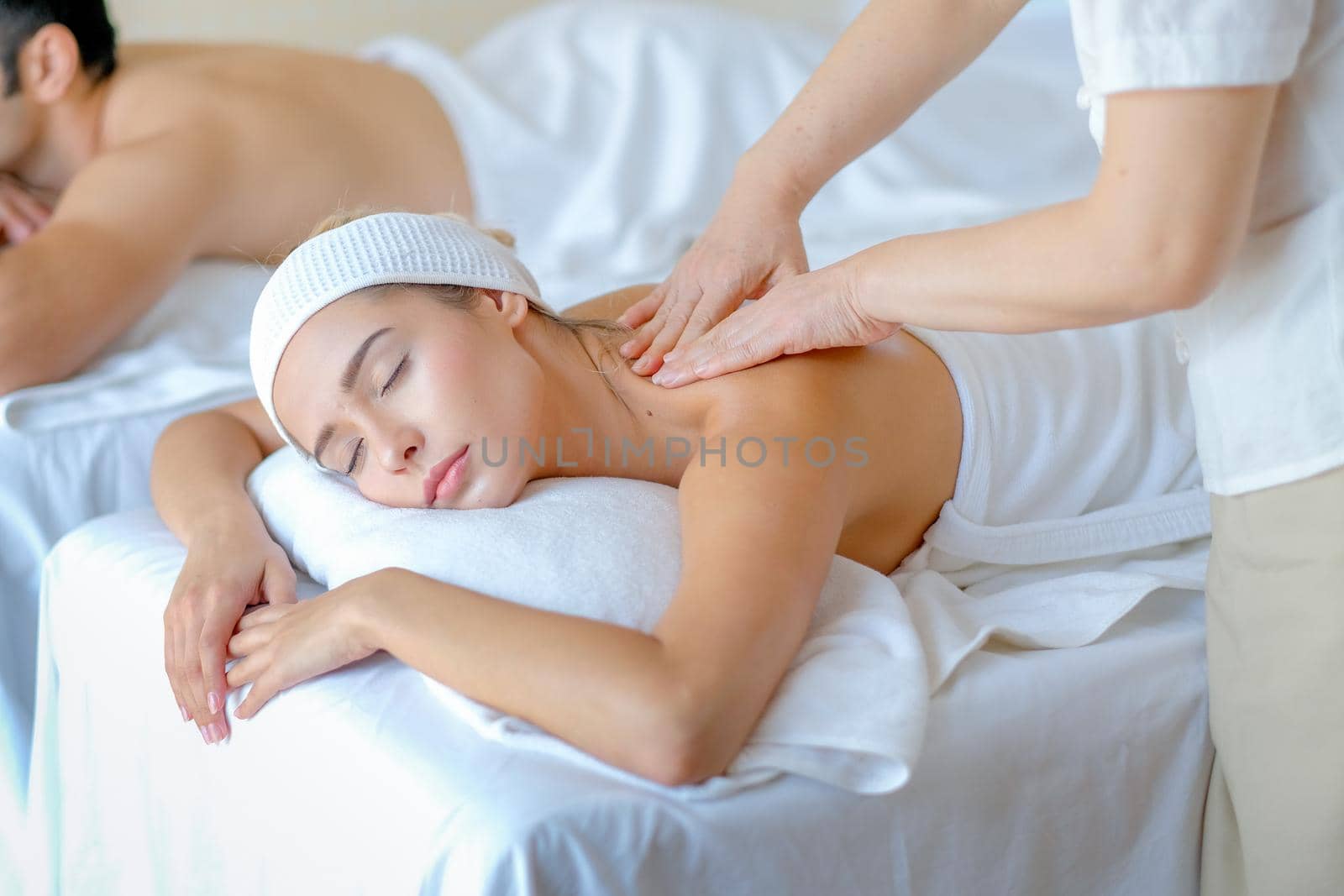 Close up of young woman enjoy with body massage in spa room with day light and the other man lie on bed beside.