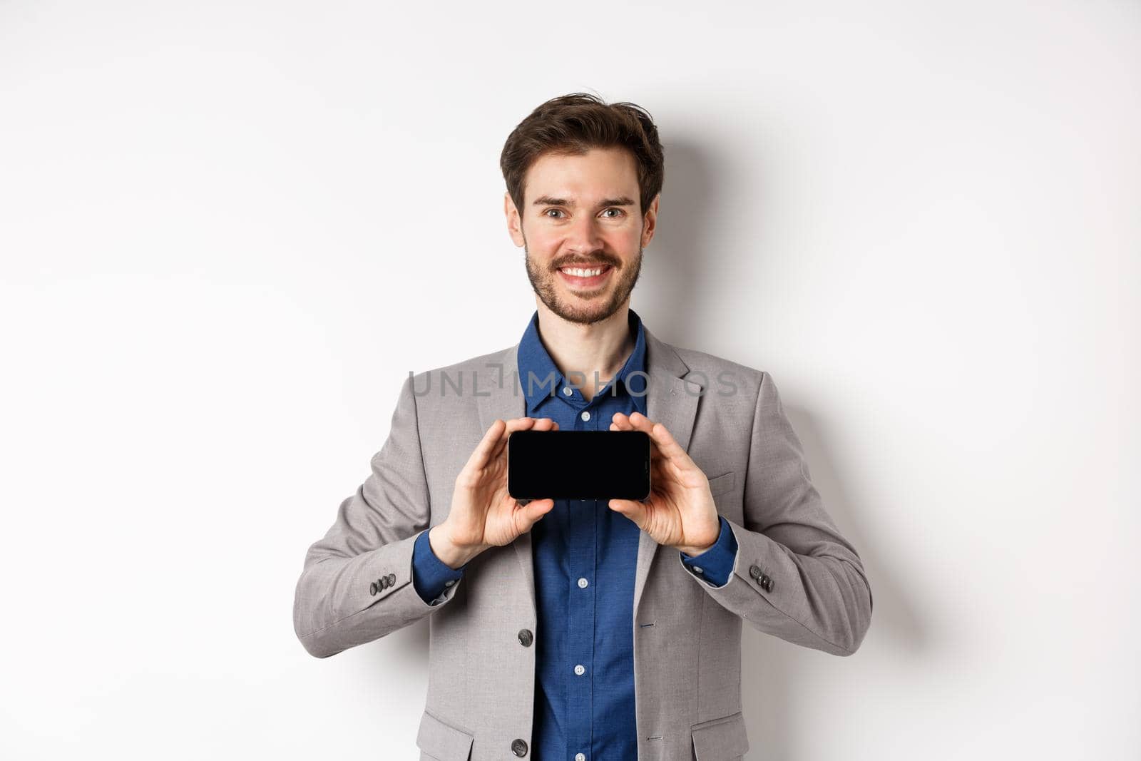 E-commerce and online shopping concept. Happy guy in business suit showing empty smartphone screen horizontally, smiling pleased at camera, white background.