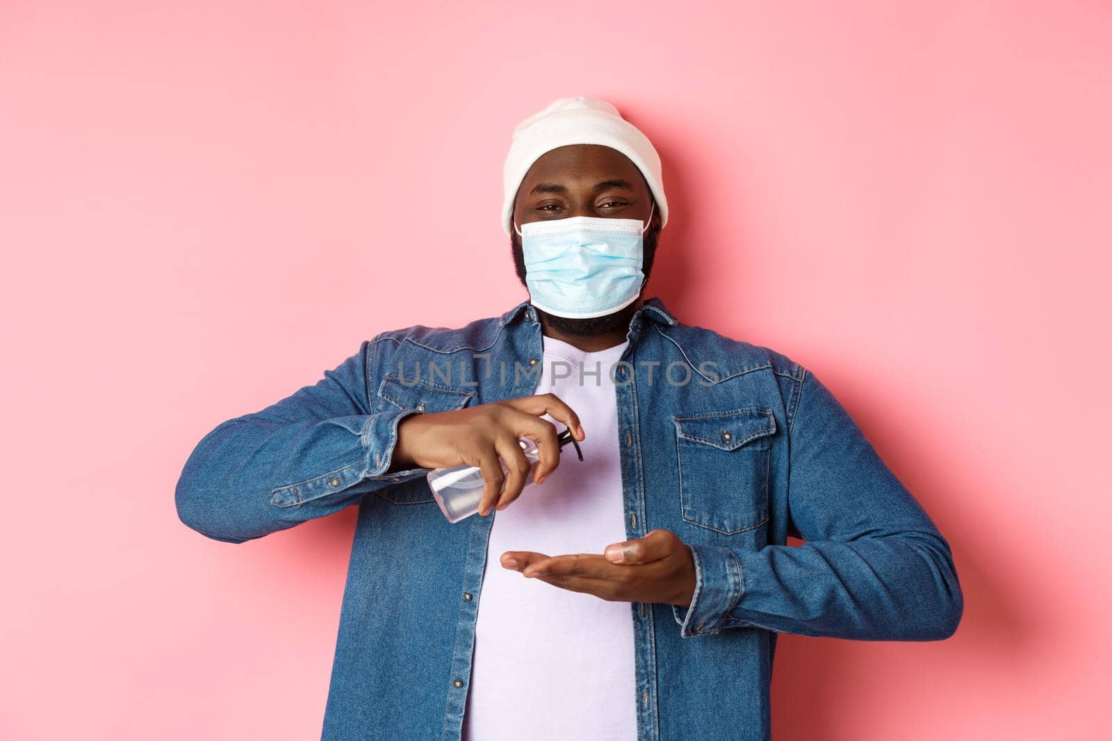 Covid-19, lifestyle and lockdown concept. Smiling african-american man in face mask cleaning hands with sanitizer, using antiseptic and looking at camera, pink background by Benzoix