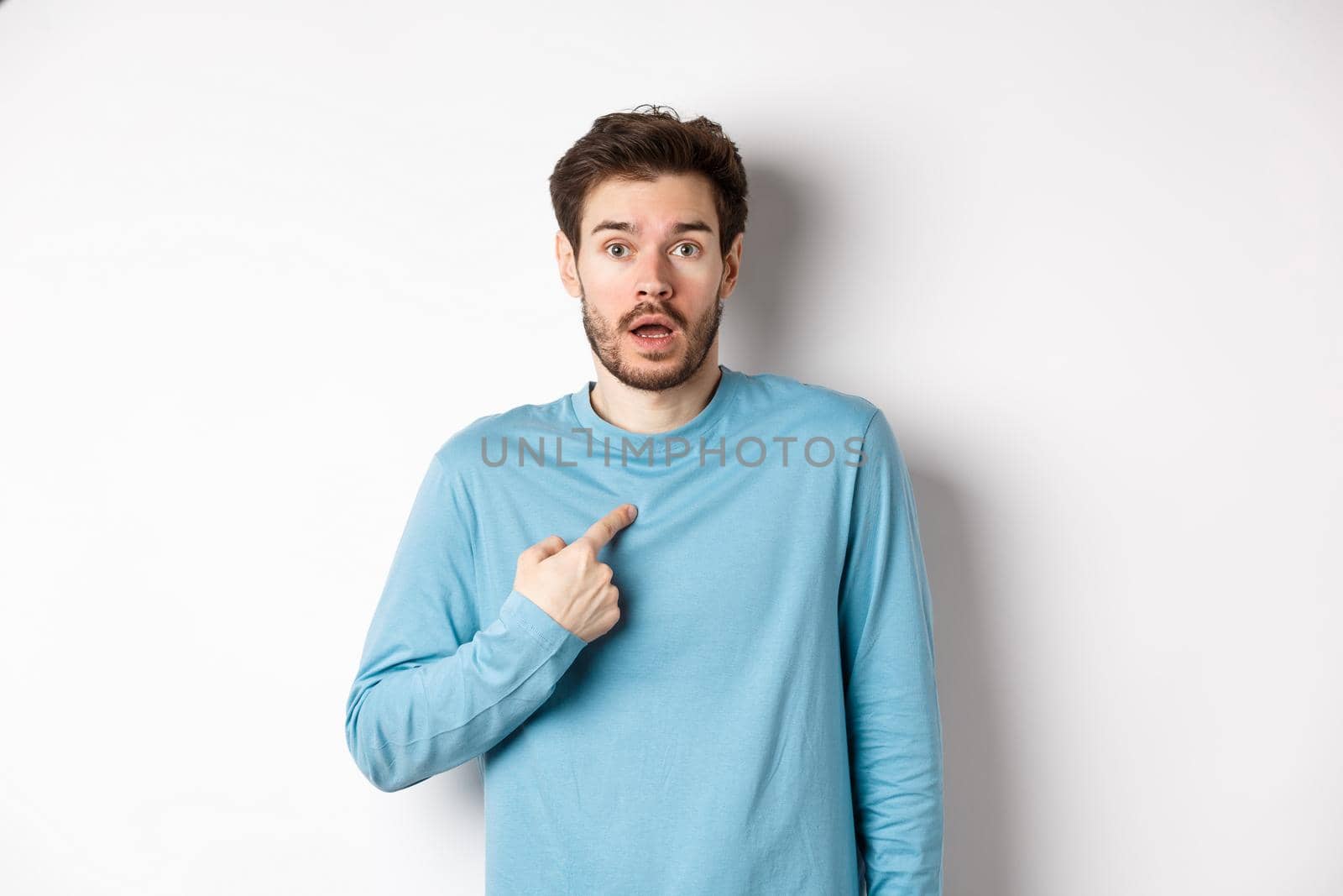 Surprised young man gasping and pointing at himself with disbelief, being chosen or named, standing over white background by Benzoix