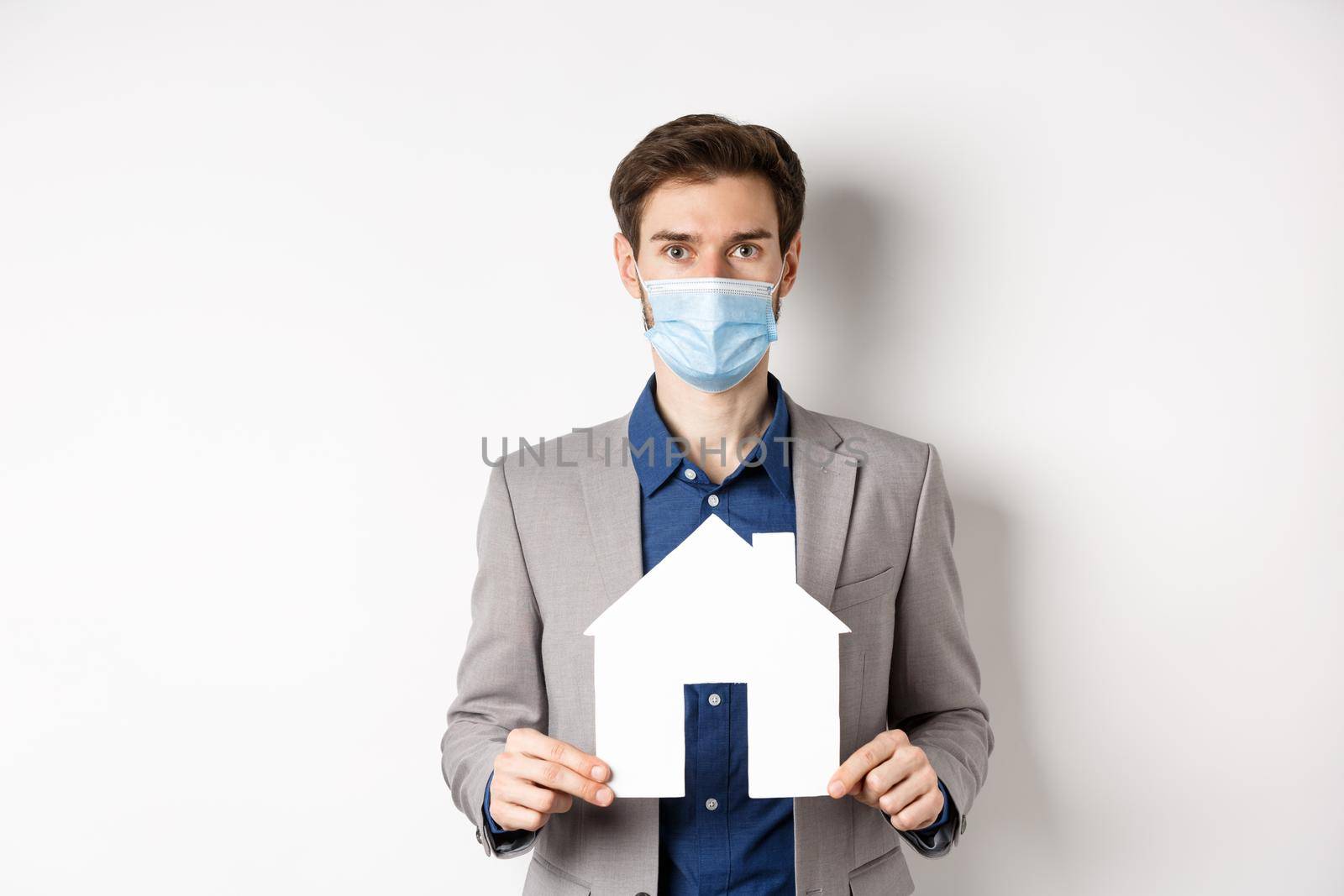 Real estate and covid-19 concept. Salesman in medical mask and suit showing paper house cutout and looking at camera, white background.