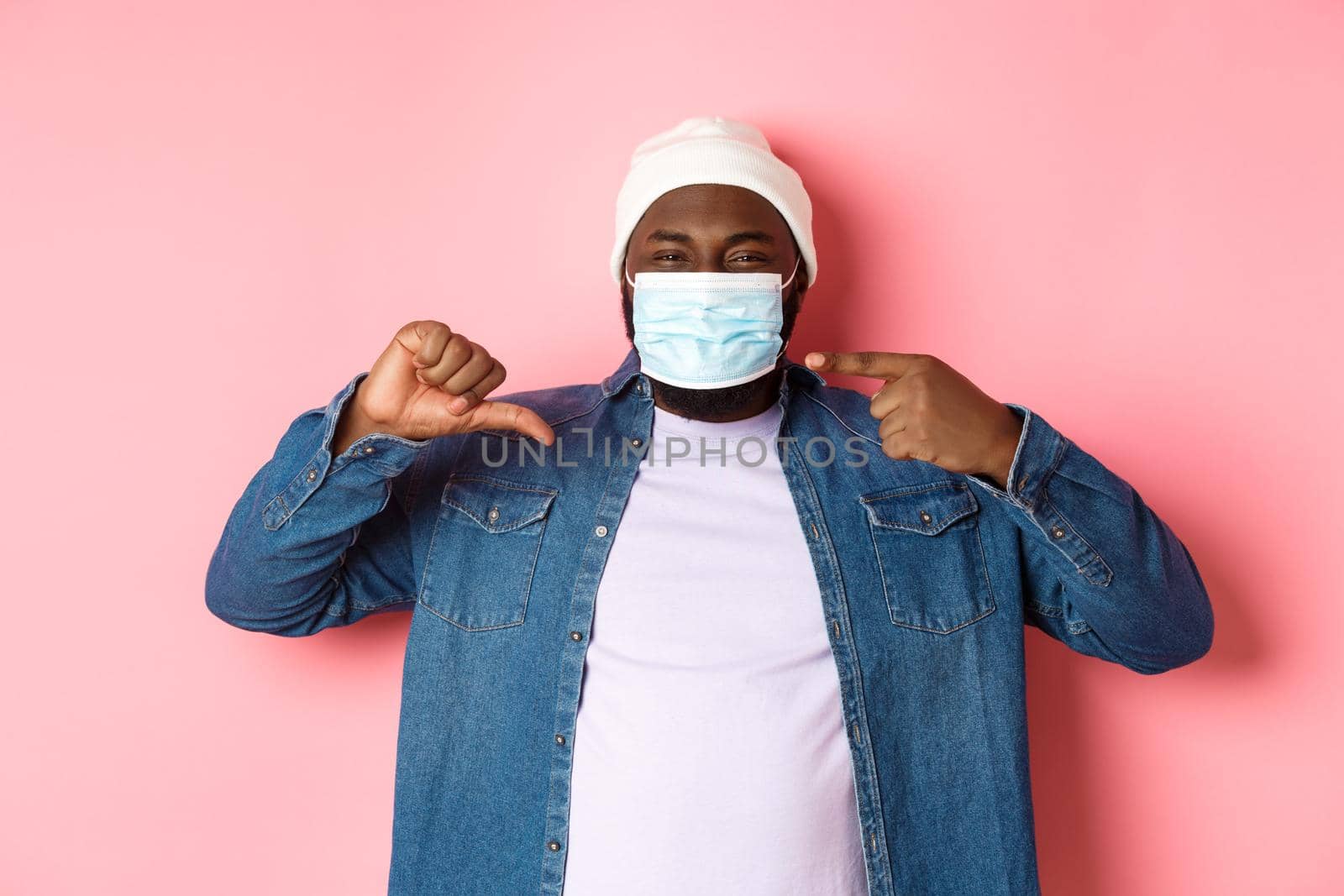 Coronavirus, lifestyle and global pandemic concept. Displeased hipster guy pointing at face mask, showing thumbs-down, dislike wearing it, pink background.