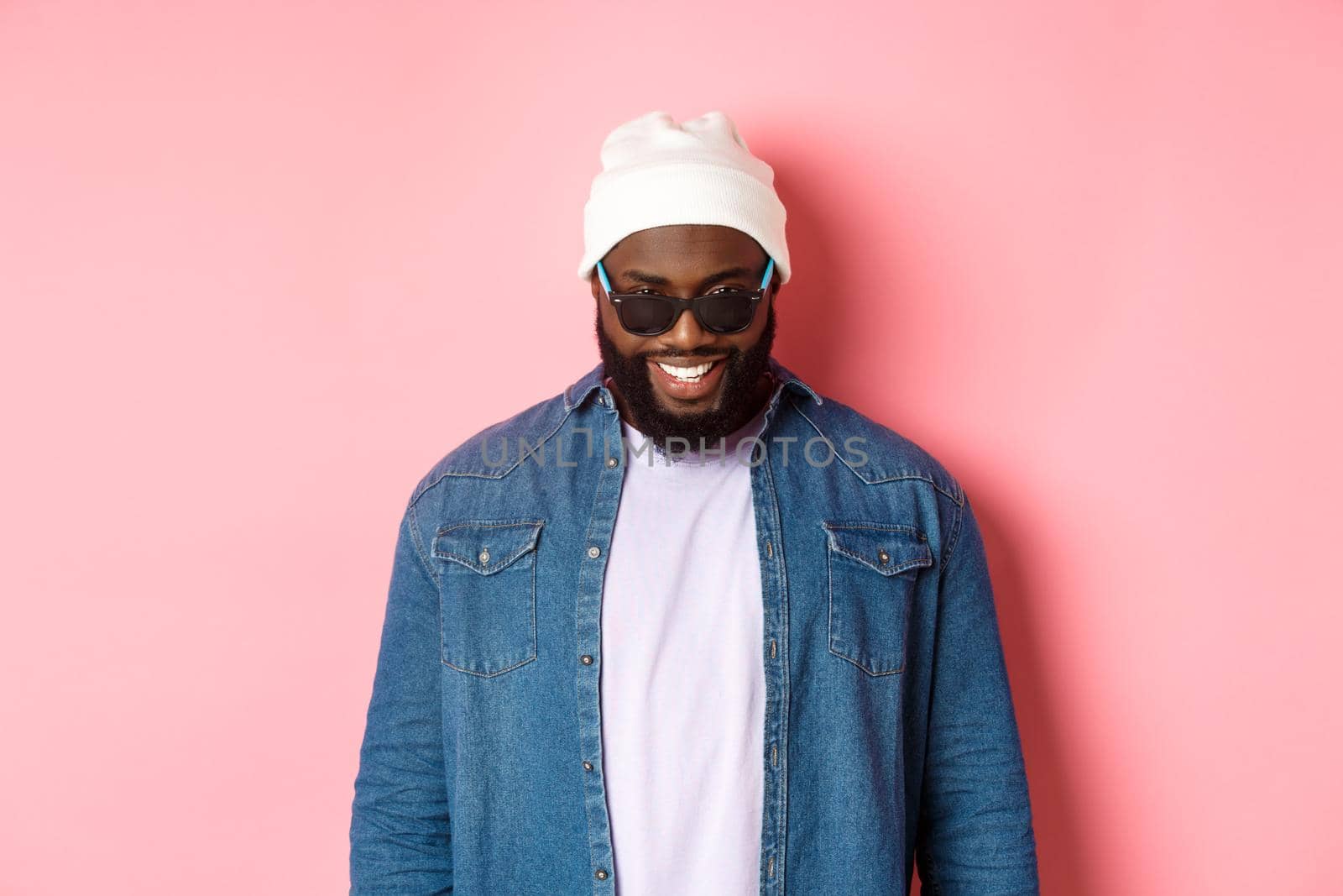Cool african-american hipster man in beanie and sunglasses, smiling pleased and looking at camera, standing confident over pink background by Benzoix