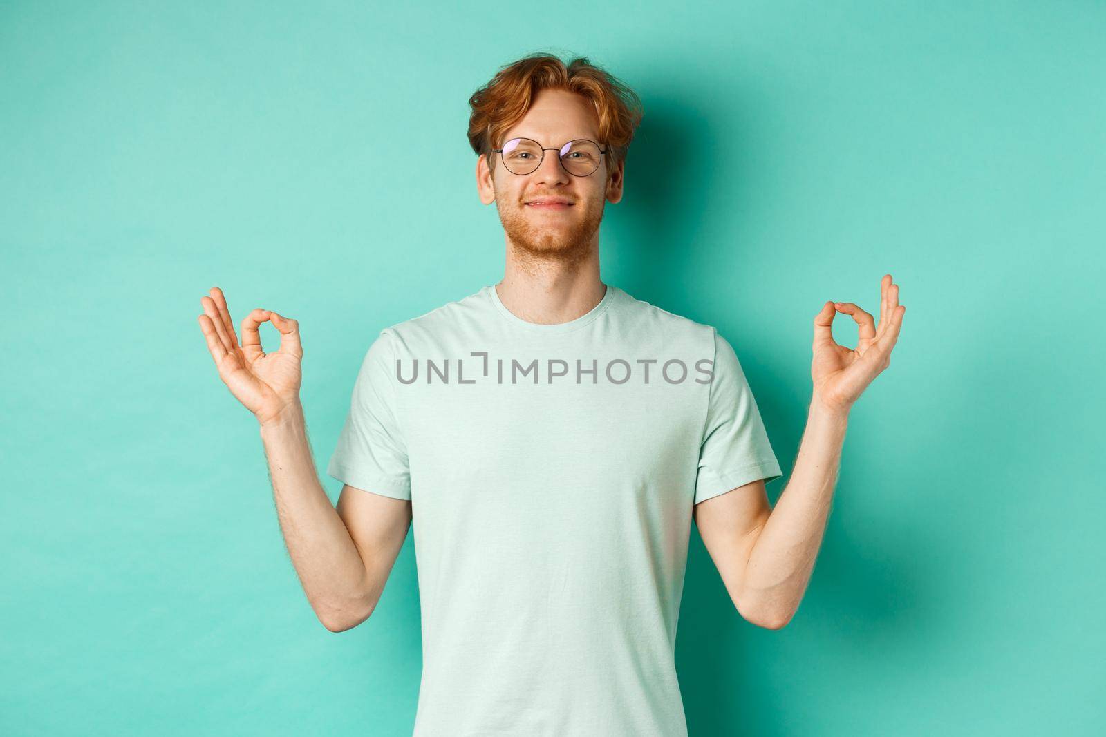 Calm and relaxed redhead man in glasses smiling, holding hands spread sideways with om mudra gesture, meditating peaceful, standing over mint background.