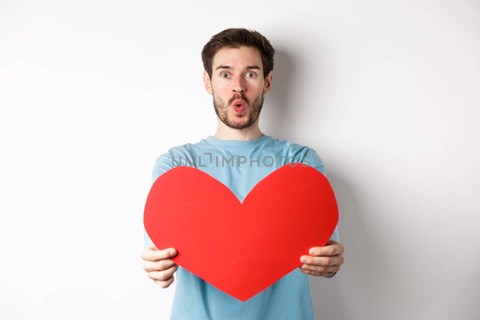 Young handsome man showing big red valentines day heart, being in love, pucker lips for kiss on valentine date, standing over white background.