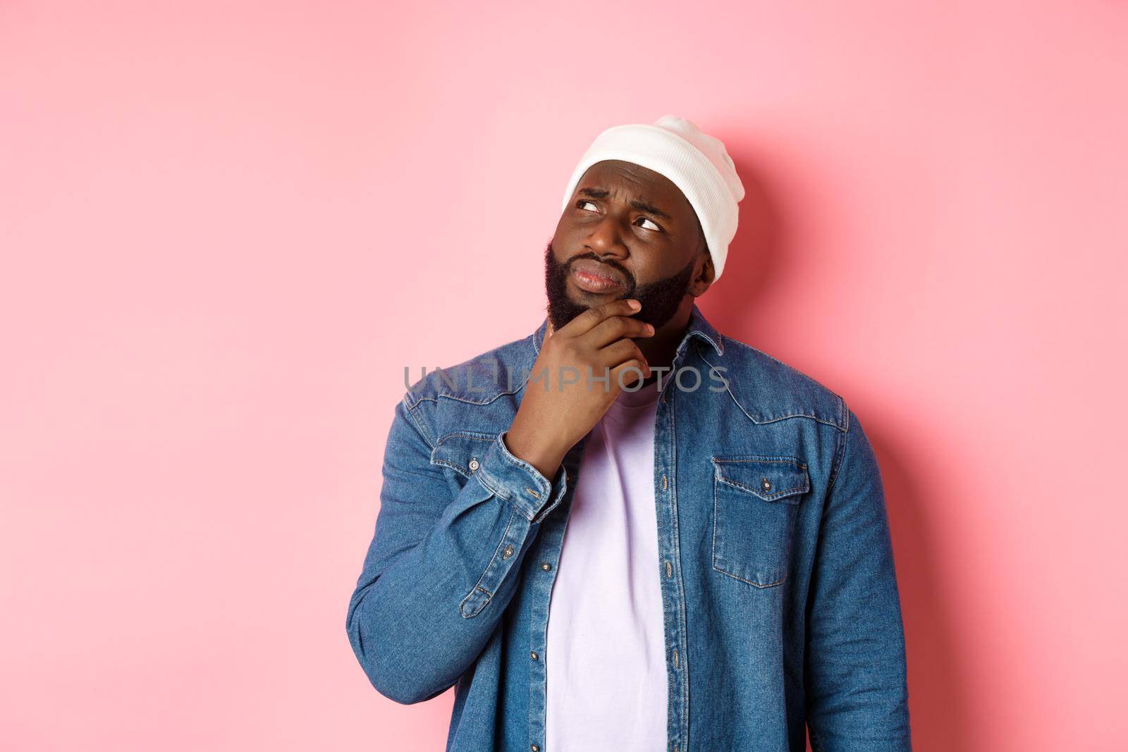 Image of puzzled and concerned black man, staring at upper left corner and thinking, making decision with doubts, standing over pink background by Benzoix