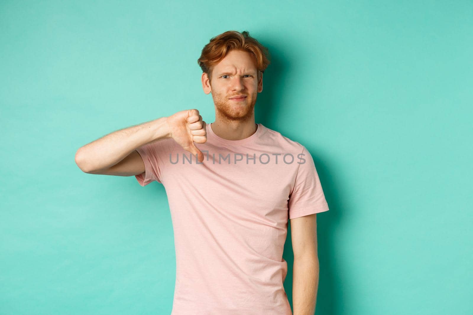 Disappointed redhead man showing thumbs-down, frowning and looking skeptical, epxress dislike and disapproval, standing over turquoise background.