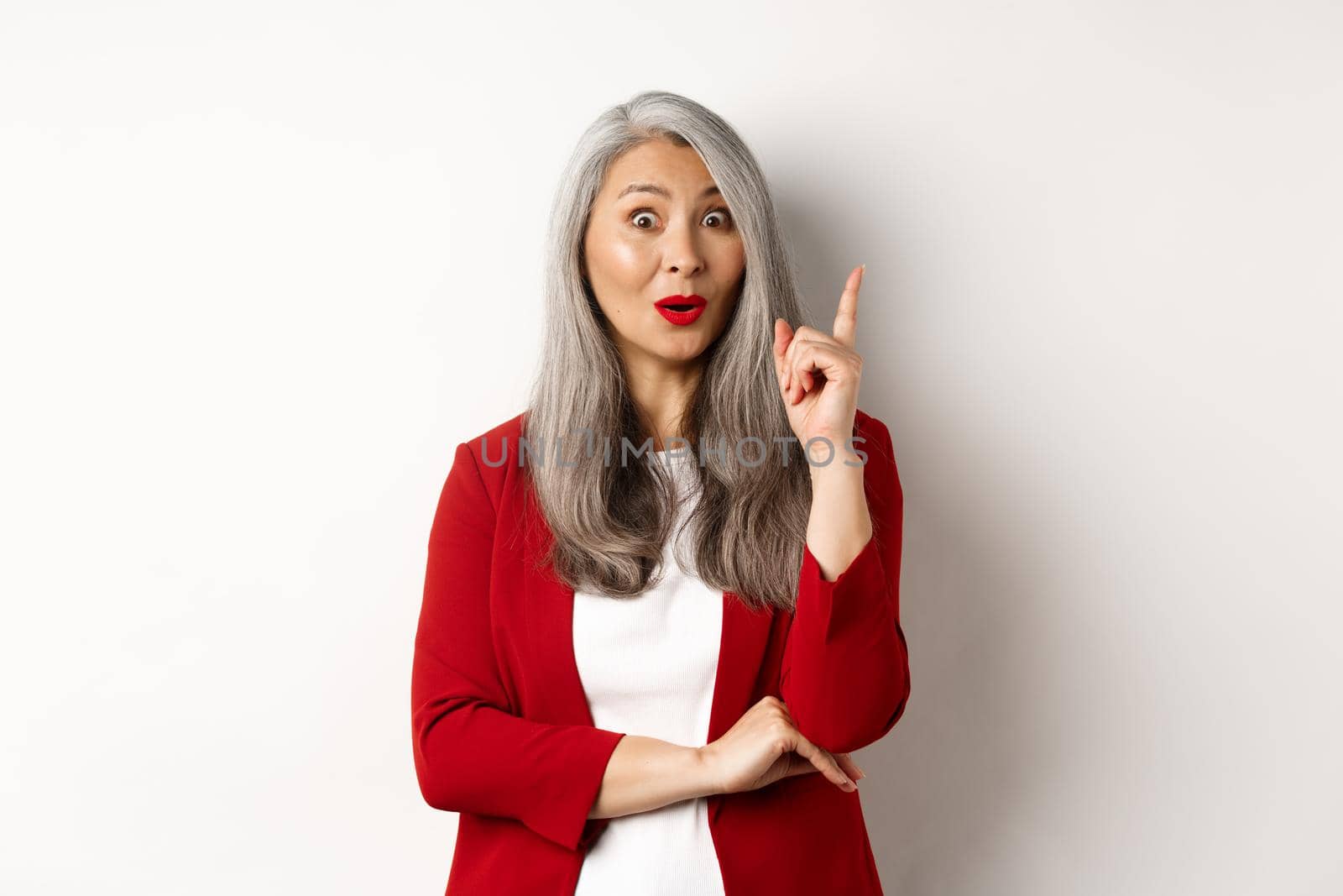 Elderly asian woman entrepreneur in red blazer having an idea, suggesting something, raising finger in eureka gesture, standing over white background.