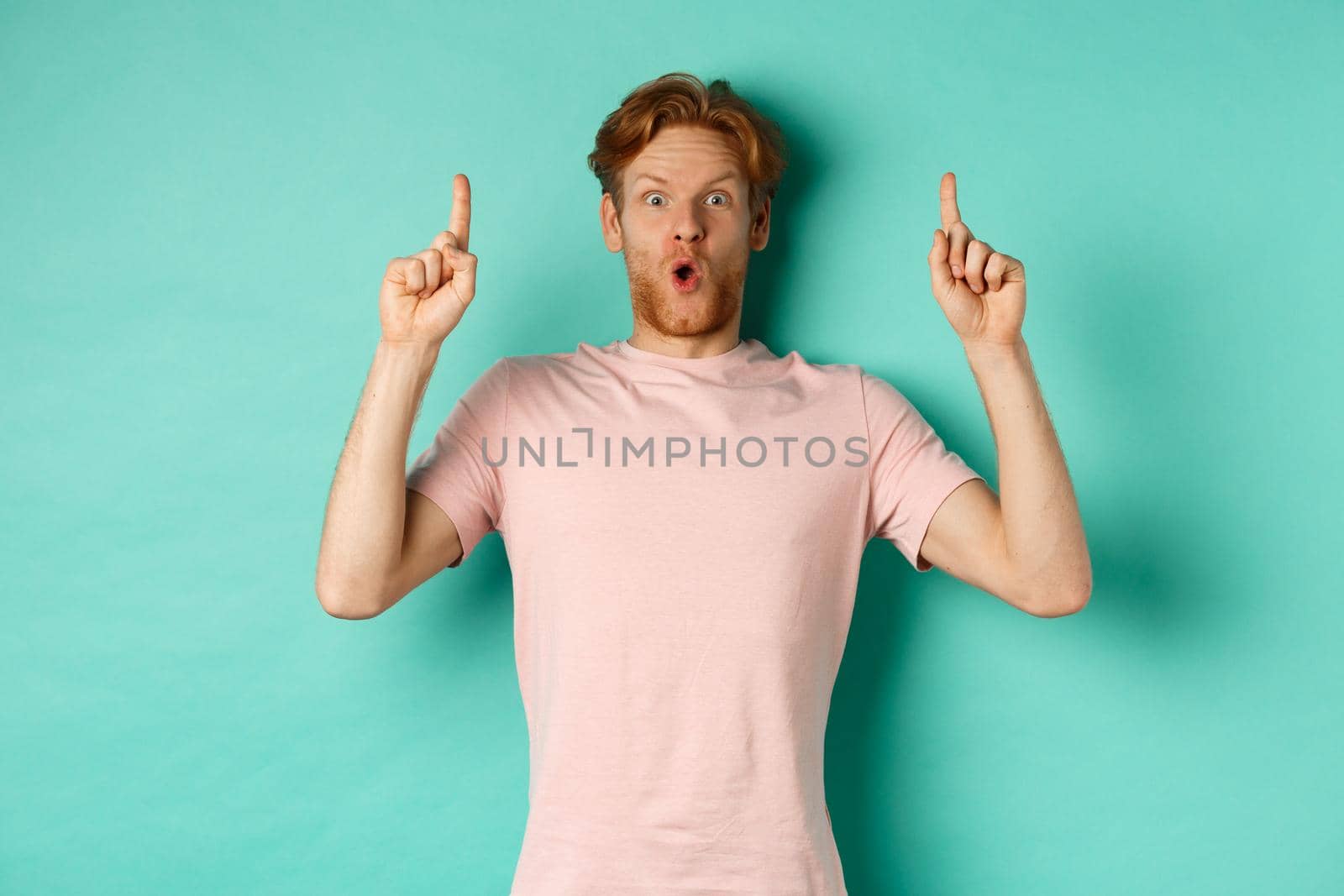 Impressed handsome guy with red hair pointing fingers up, demonstrate promo offer, standing in t-shirt over mint background by Benzoix