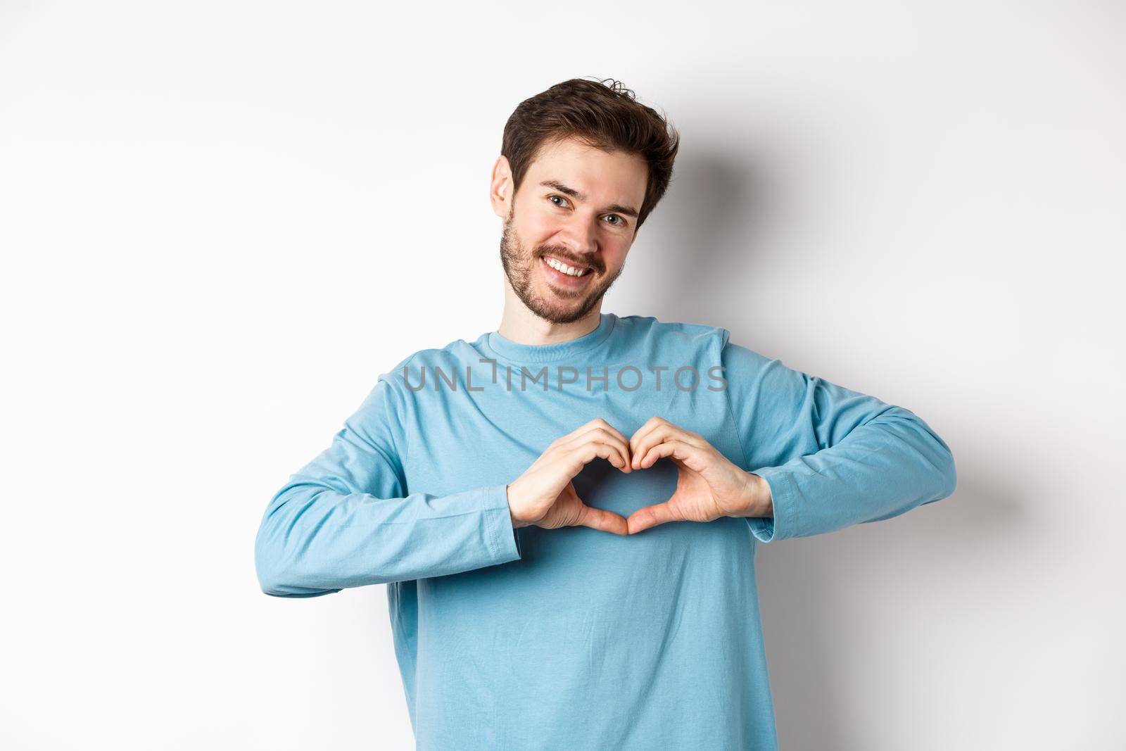 Handsome boyfriend saying I love you, showing heart gesture and smiling at camera, express love and romantic feeling, standing over white background.