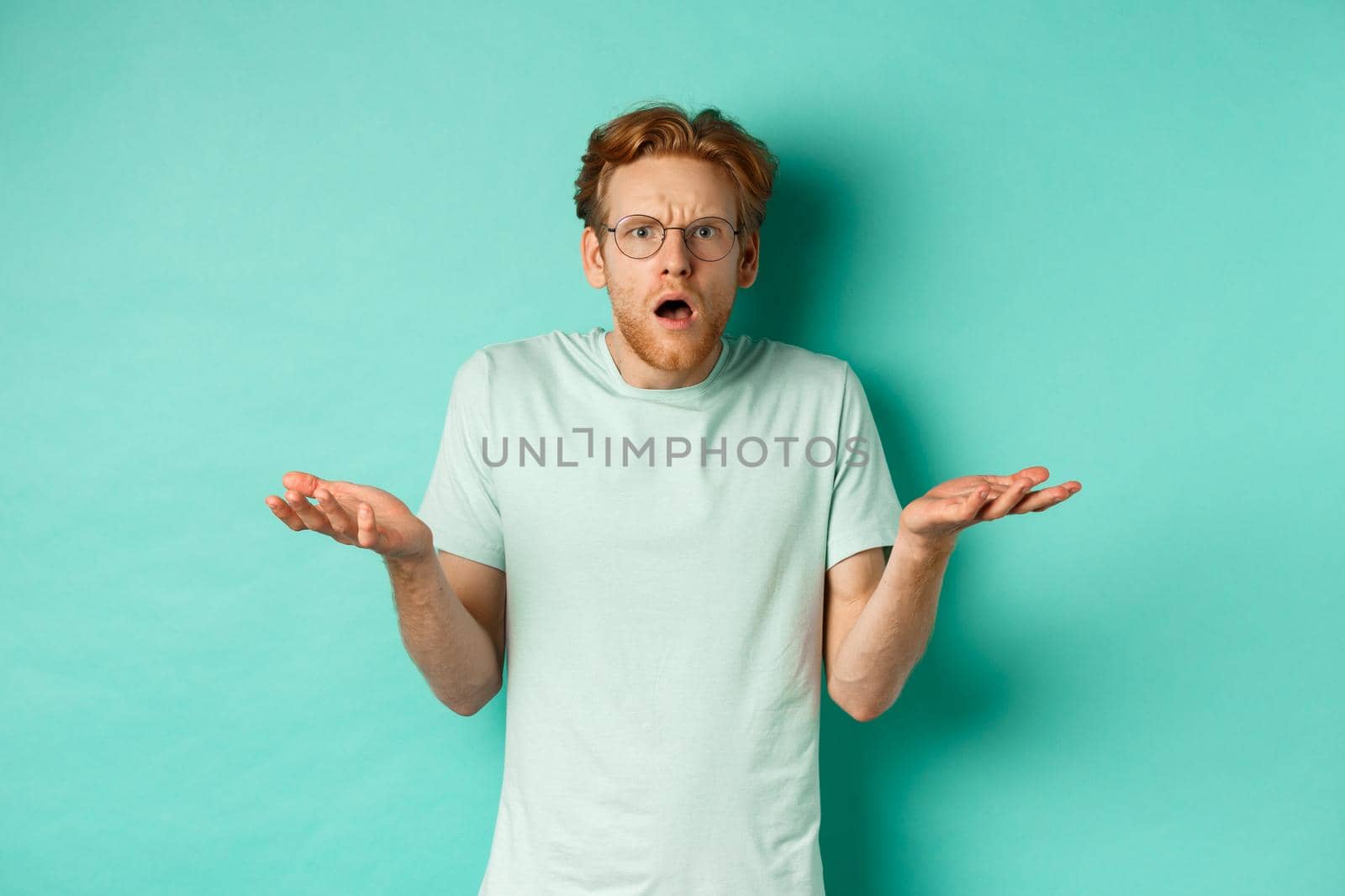 Confused and shocked guy with red messy hair and glasses, shrugging and raising hands, staring at something strange, cant understand, standing over turquoise background by Benzoix