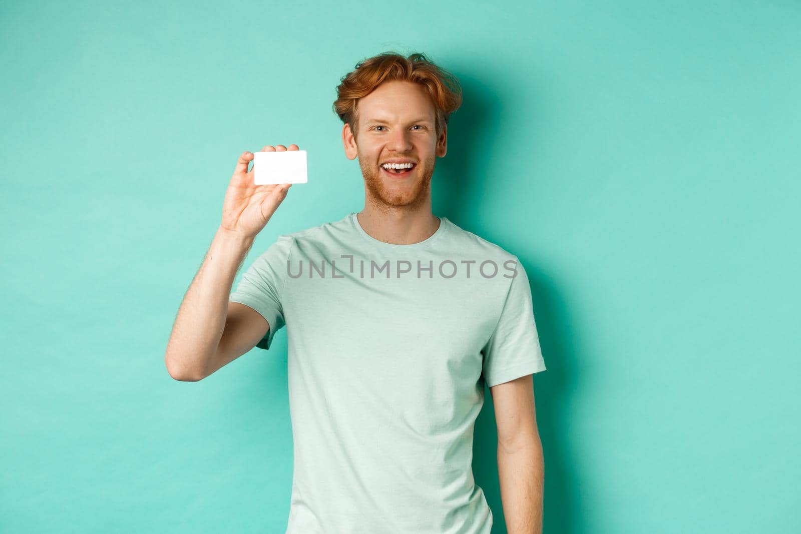 Shopping concept. Cheerful young man in t-shirt showing plastic credit card and smiling, standing over mint background by Benzoix