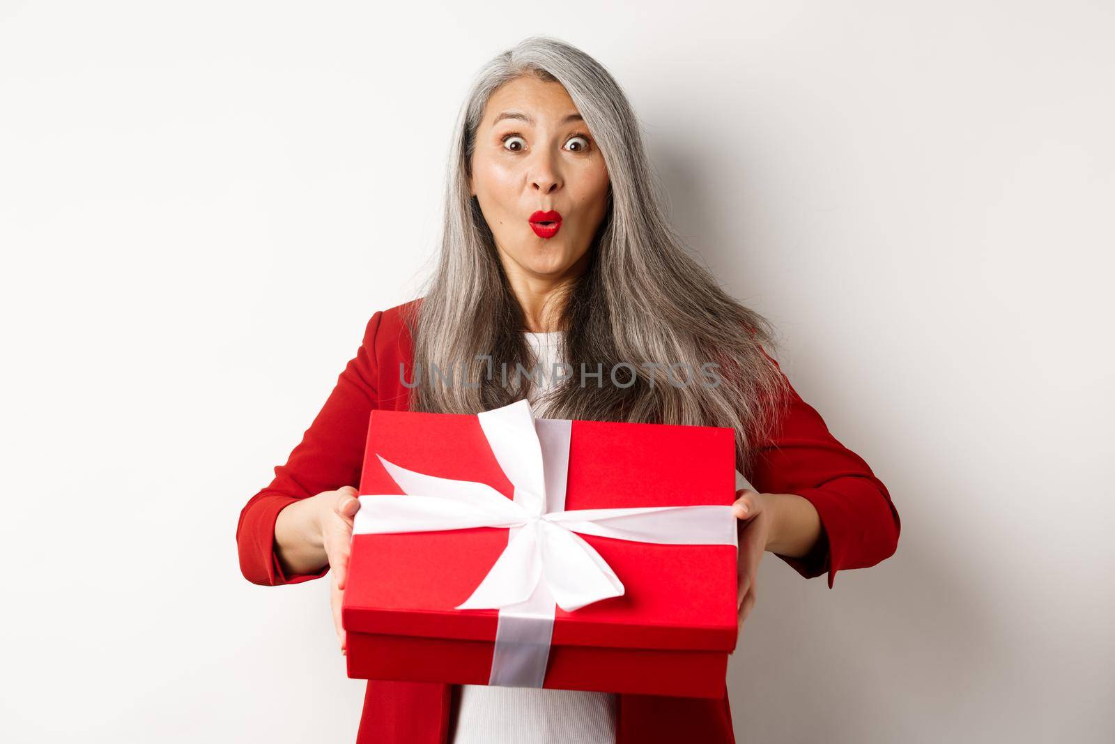 Surprised asian senior woman receiving present on mother day, holding red box with gift and looking amazed at camera, white background by Benzoix