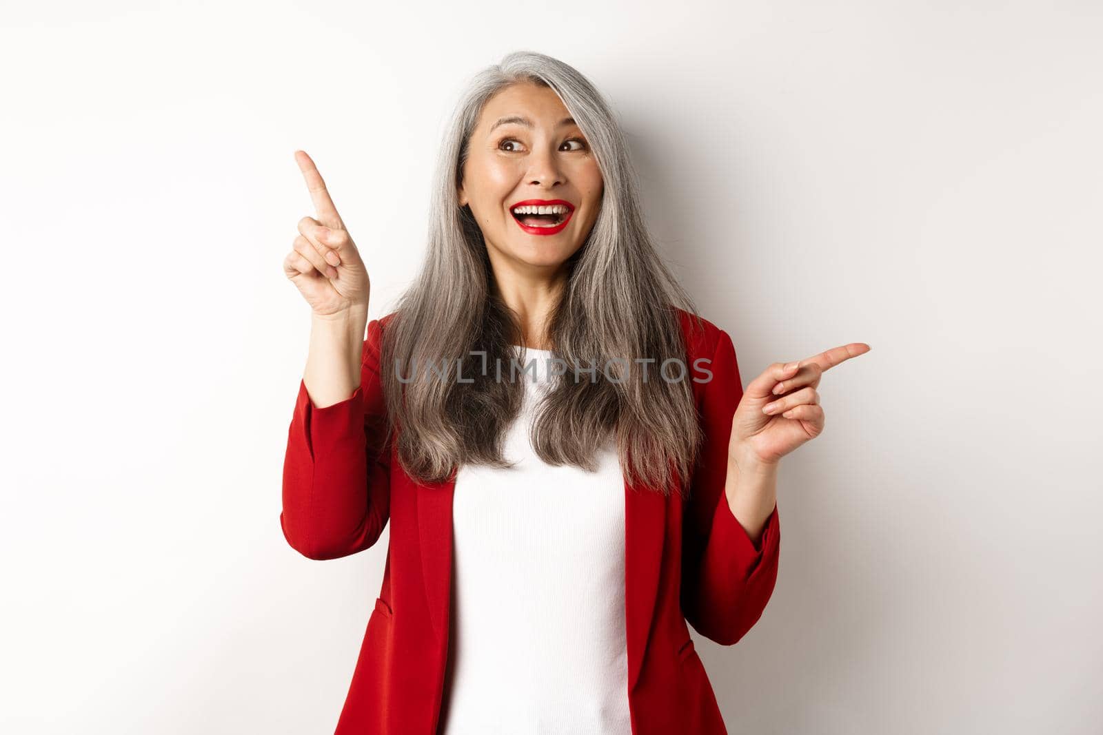 Cheerful asian lady smiling, pointing fingers sideways at two promo offers, standing happy in red blazer over white background by Benzoix