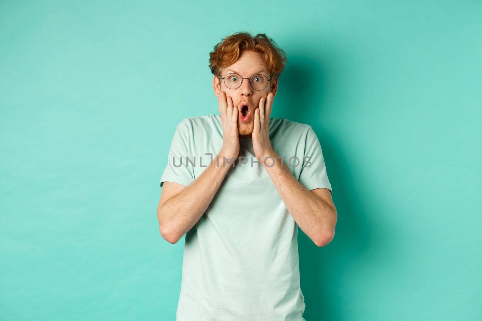 Shocked redhead guy in glasses checking out awesome promotion offer, gasping amazed and holding hands on face, staring at camera, mint background.