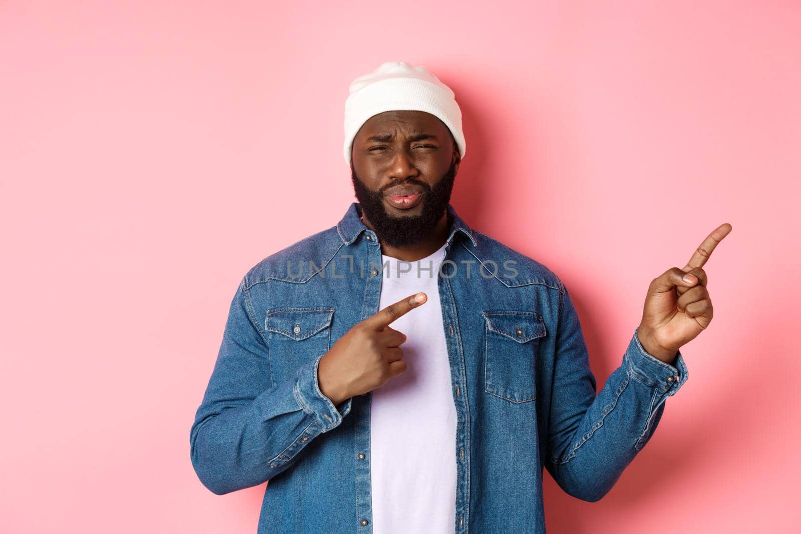 Disappointed and skeptical african-american guy complaining, cringe from disgust, frowning and grimacing from dissatisfaction, pointing fingers right, pink background by Benzoix