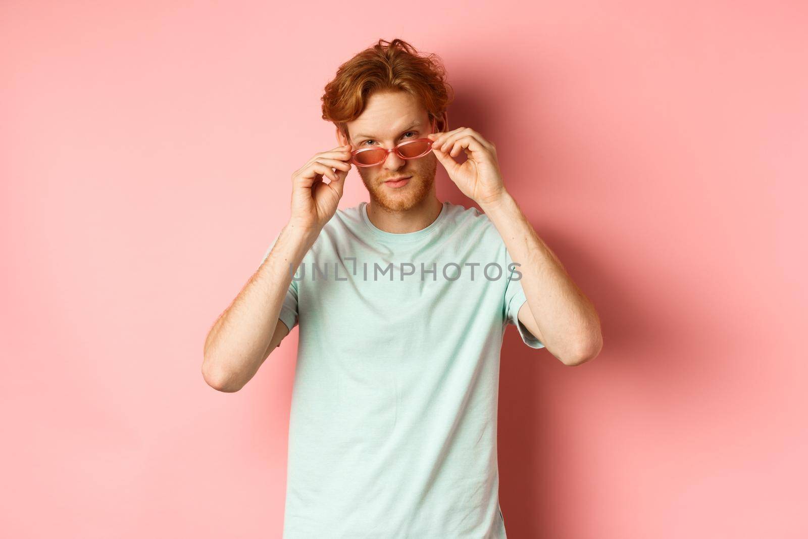 Handsome redhead man in summer sunglasses looking sassy at camera, standing over pink background by Benzoix