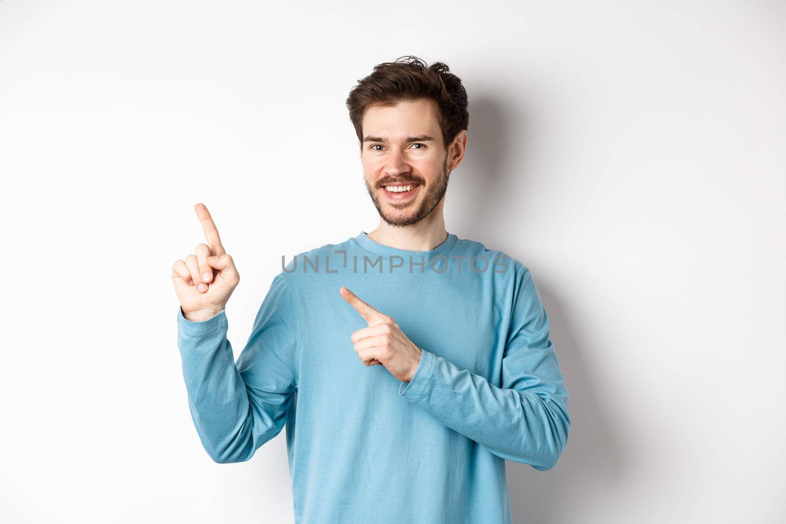 Cheerful bearded man showing advertisement on copy space, pointing fingers at upper left corner logo and smiling, standing over white background by Benzoix