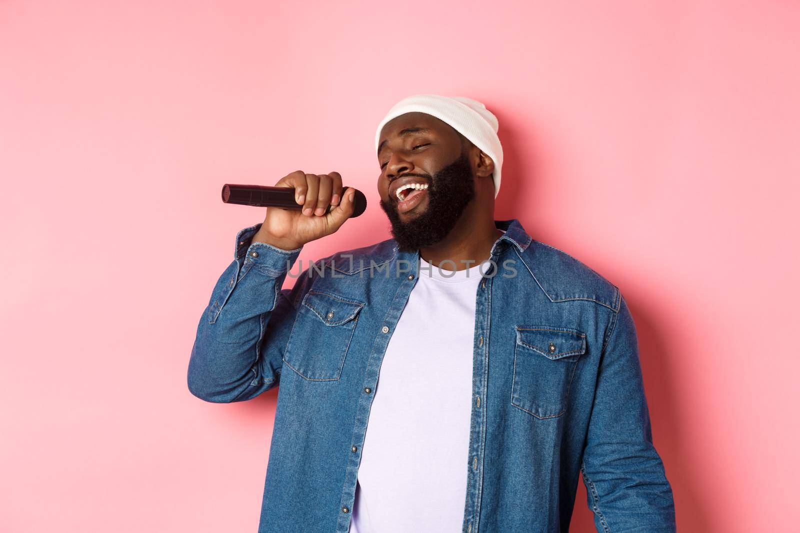 Handsome Black man in beanie and denim shirt singing karaoke, holding microphone, standing over pink background by Benzoix