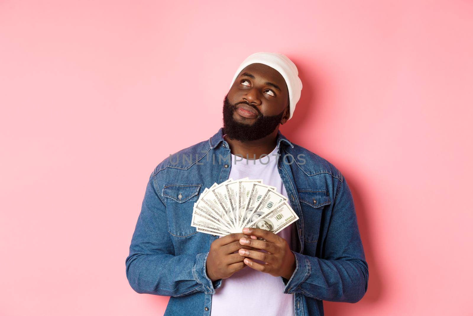 Dreamy african-american man thinking about shopping, holding money dollars and looking upper left corner, standing over pink background.