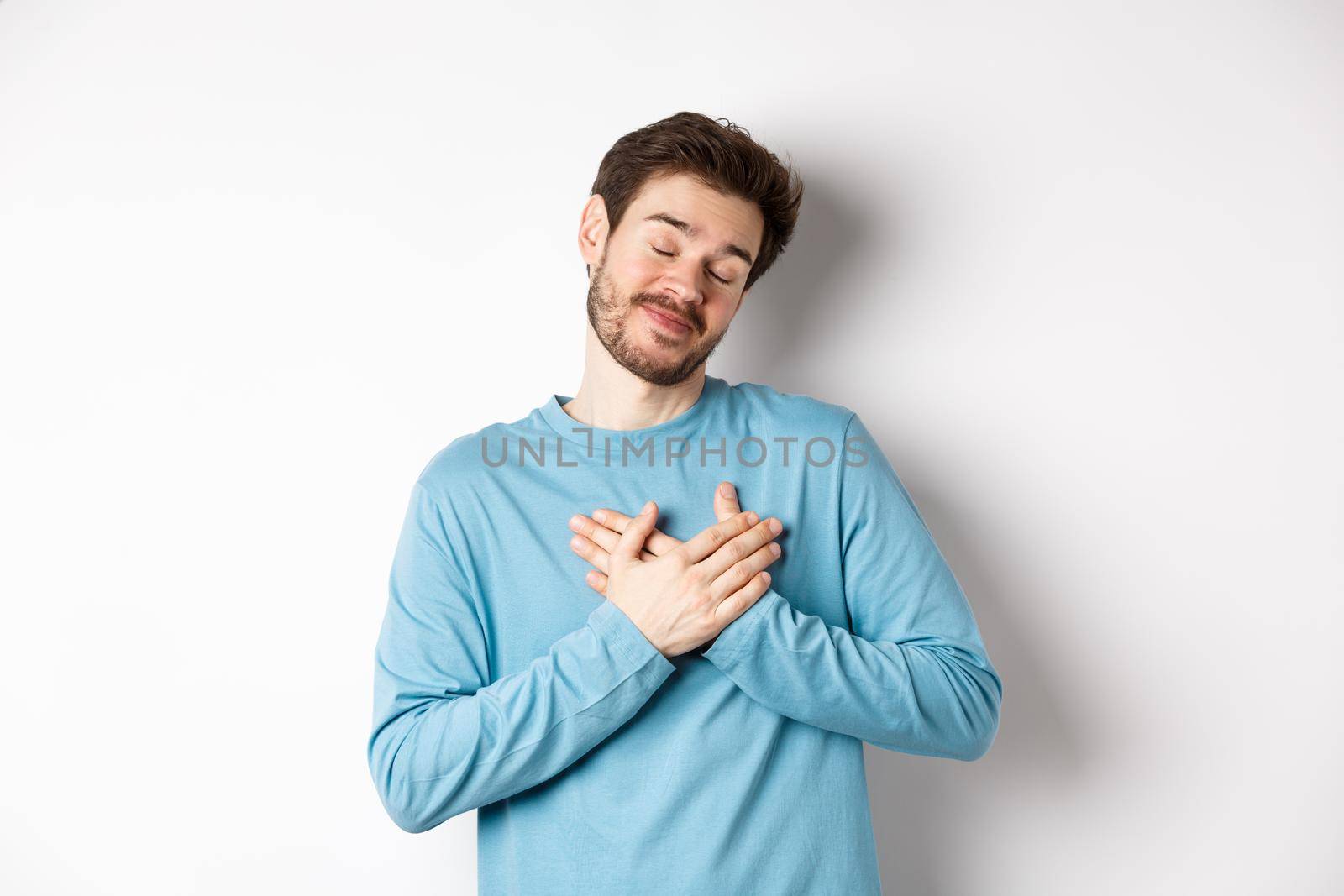 Handsome young man feeling romantic, holding hands on heart and thinking about good pleasant memories, standing over white background.