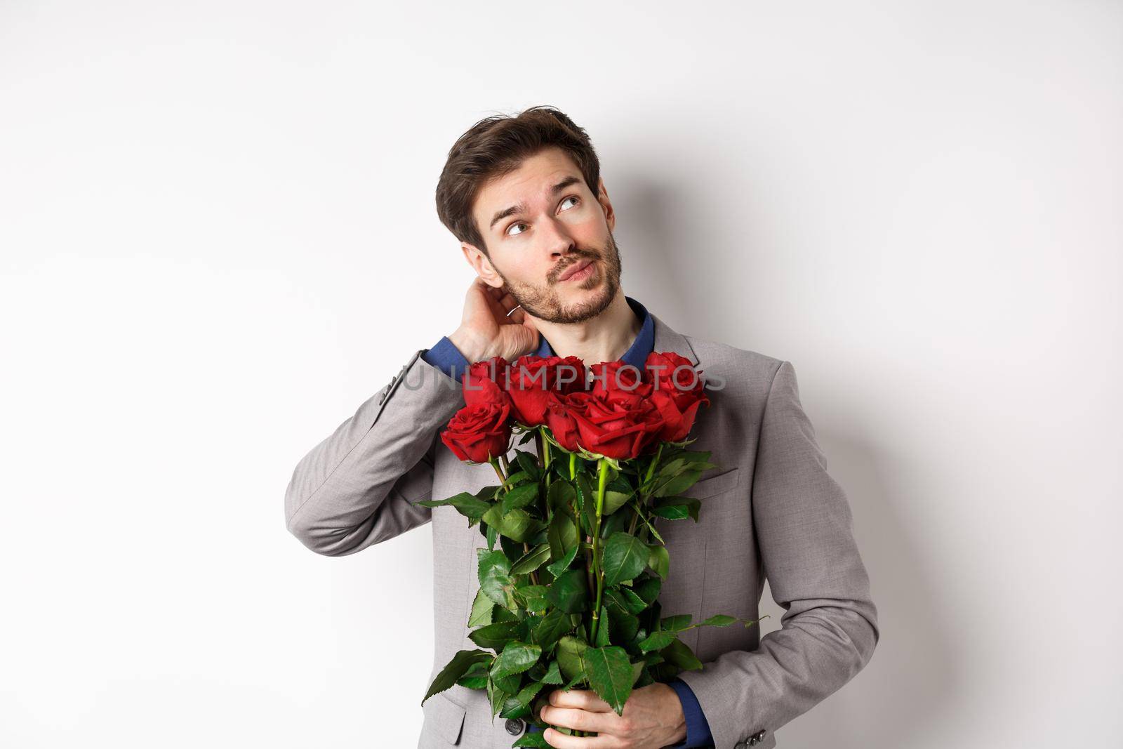 Thoughtful young man in suit going on valentines day date, holding bouquet of roses, thinking and scratching head, standing against white background.