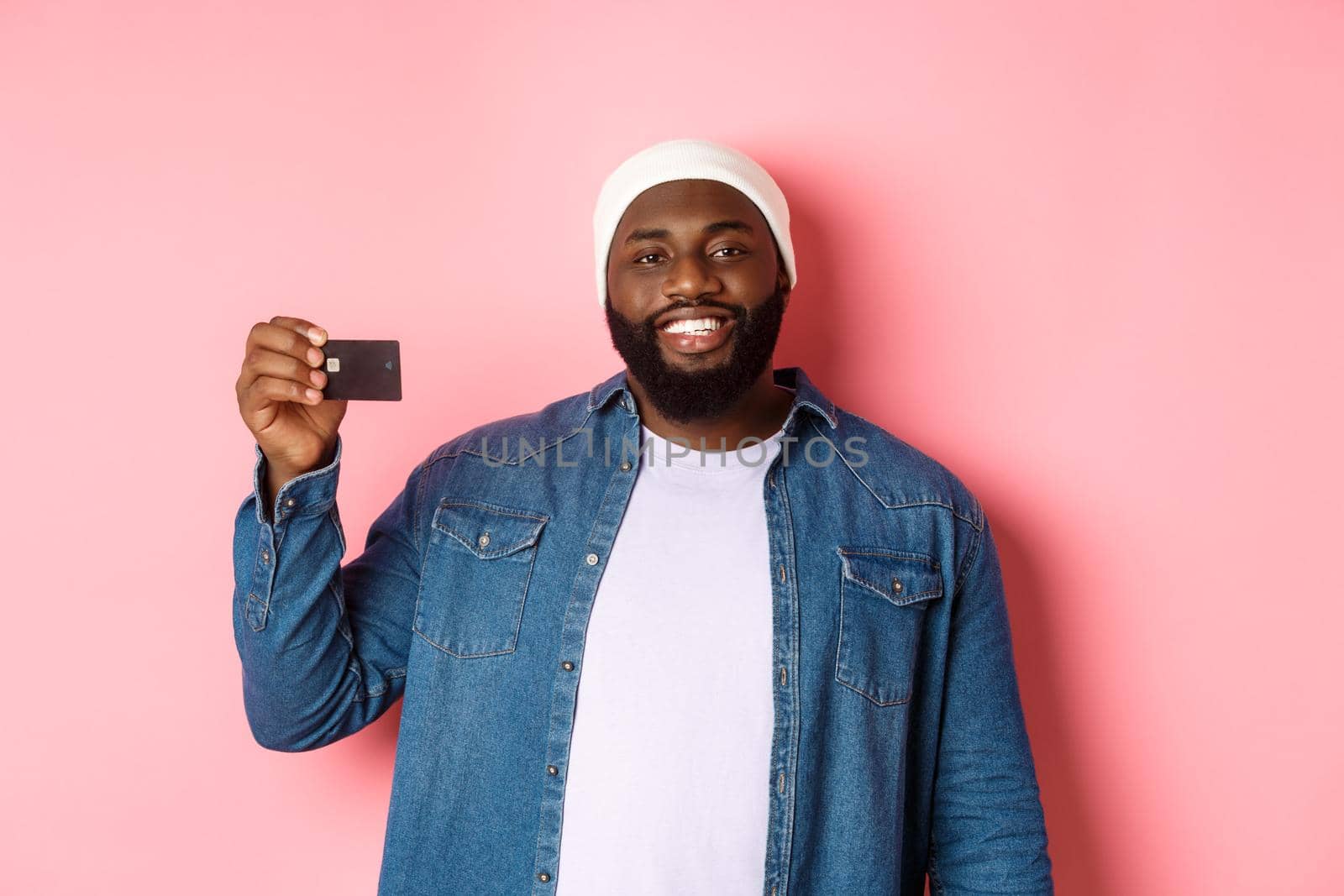 Shopping concept. Satisfied young bearded man in beanie showing credit card, smiling pleased, making purchase, standing over pink background.