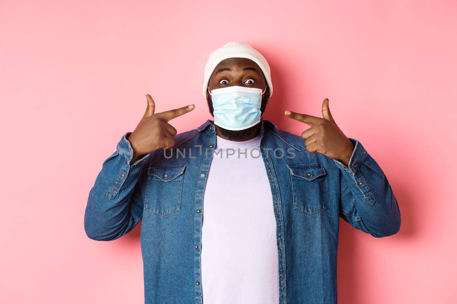 Coronavirus, lifestyle and social distancing concept. Happy Black man in beanie pointing at his face mask, smiling at camera, standing over pink background.