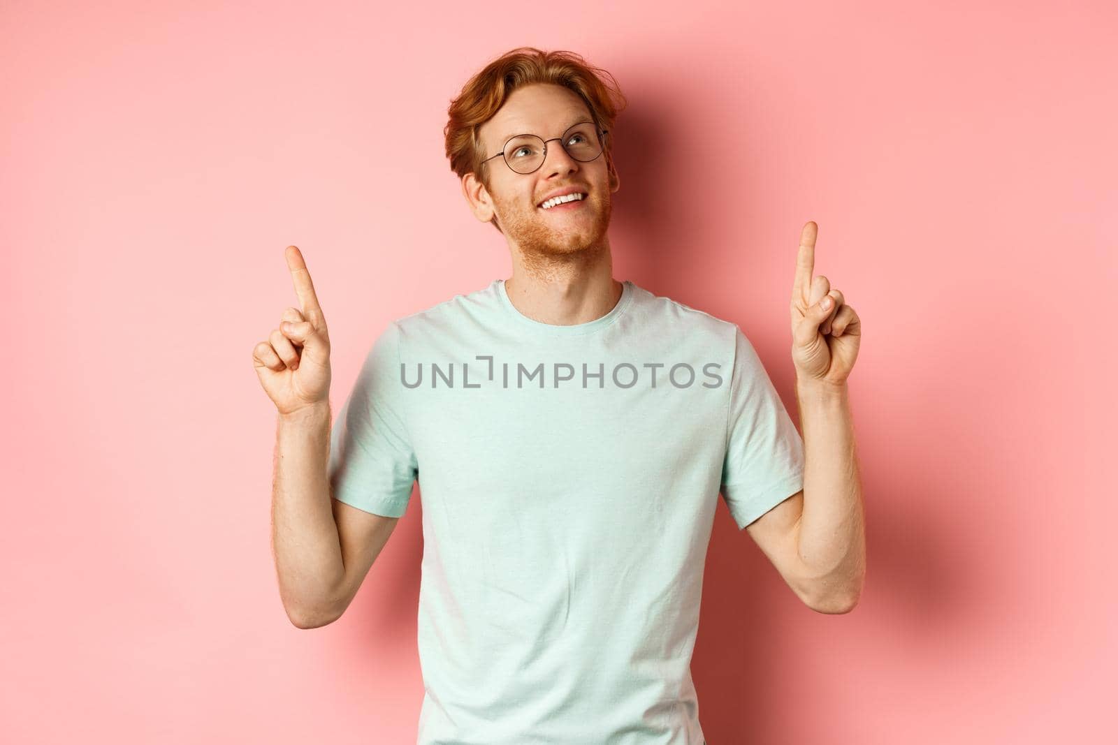 Dreamy young man with red hair and beard pointing fingers and looking cheerful up, admire something, standing over pink background by Benzoix