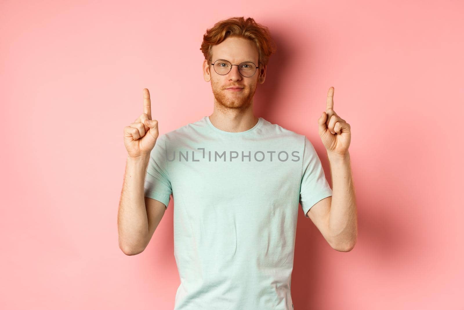 Confident and pleased young man with red hair, wearing glasses and t-shirt, pointing fingers up and smiling with smug face, showing good deal, standing over pink background.